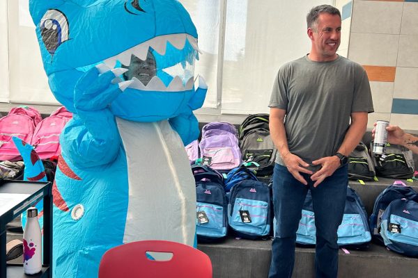 UCLI Board member, Jascha Clark passes out backpacks and Meadowlark school counselor  in an inflatable dinosaur costume greeting families at Meadowlark Elementary.